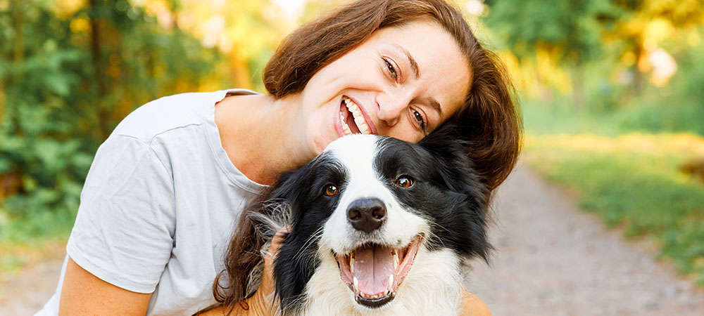 Eine junge Frau und ihr Border Collie Welpe lachen an einem Sommertag strahlend in die Kamera